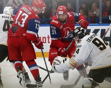 Хоккейный матч: два игрока в красной форме команды с логотипом Локомотив атакуют ворота. Голкипер в белой форме команды соперника защищает шайбу. На заднем плане видны зрители
